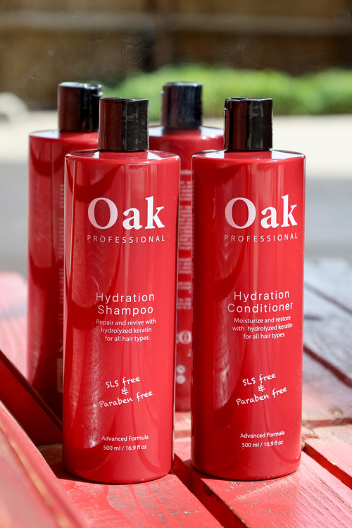 Red bottles of Oak Professional Hydration Shampoo and Conditioner on a wooden surface with a blurred outdoor background.