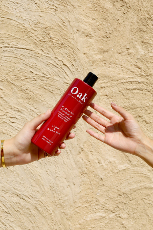 Two hands against a textured beige wall, one hand holding a red Oak Professional Hydration Shampoo bottle while the other reaches toward it, showcasing the product.
